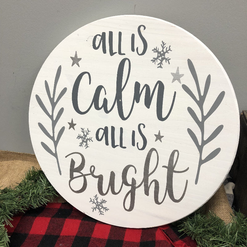 Round wooden sign with 'All is Calm, All is Bright' text and winter elements on a gray background.