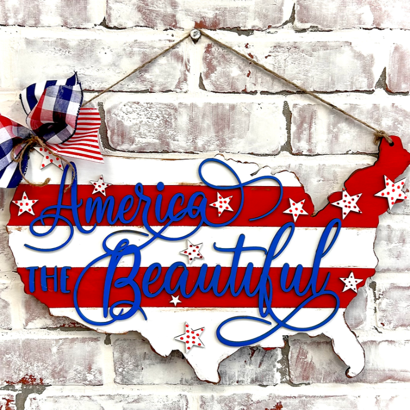 A door hanger in the shape of the United States with 'America the Beautiful' written in cursive, decorated with red and blue stripes and white stars, attached to a white brick wall.
