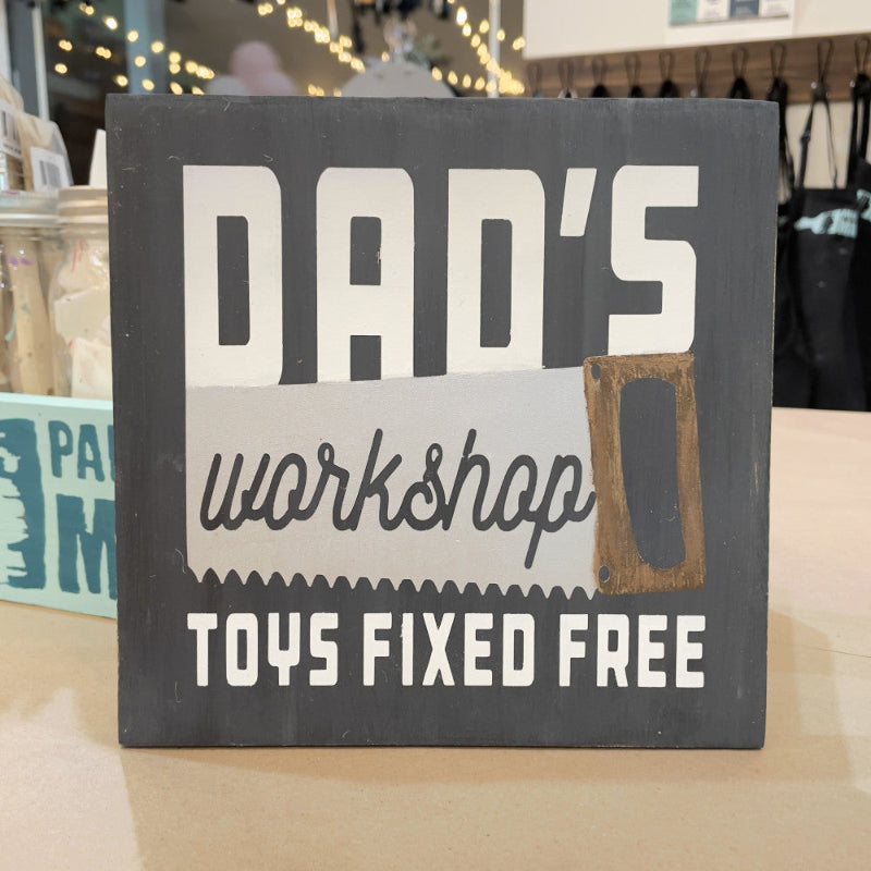 A square decorative sign with the text 'DAD'S workshop TOYS FIXED FREE' in various colors, mounted on a display shelf.