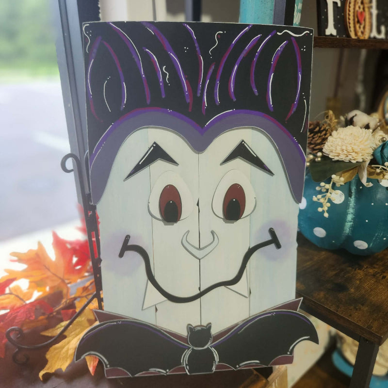 A Halloween decorative sign featuring a stylized vampire face with purple hair, large red eyes, and a bat at the bottom, displayed on a shelf with autumn leaves and a teal pumpkin in the background.