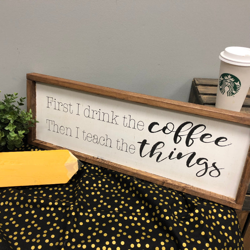A wooden plank sign with the text 'FIRST I DRINK THE COFFEE THEN I TEACH THE THINGS' written on it, accompanied by a Starbucks cup and a yellow object, likely a staple, on a table with a polka-dot patterned fabric in the background.