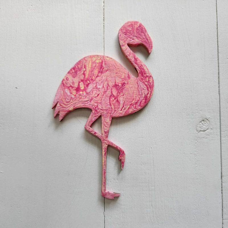 A pink flamingo wooden cutout with a paint pour design, placed against a white wooden background.