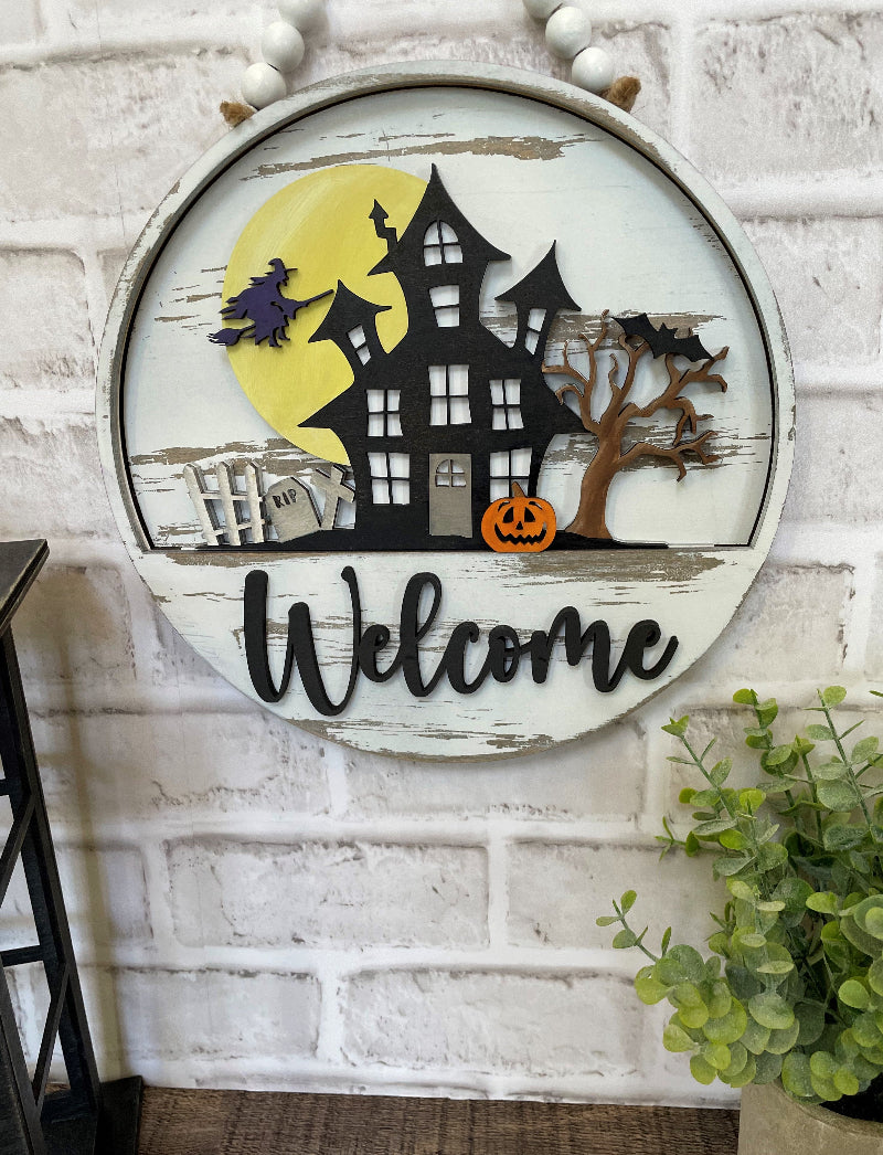 Haunted House interchangeable wooden Welcome sign insert featuring a black haunted house, yellow full moon, flying witch, graveyard, bats, spooky tree and orange jack-o'-lantern.