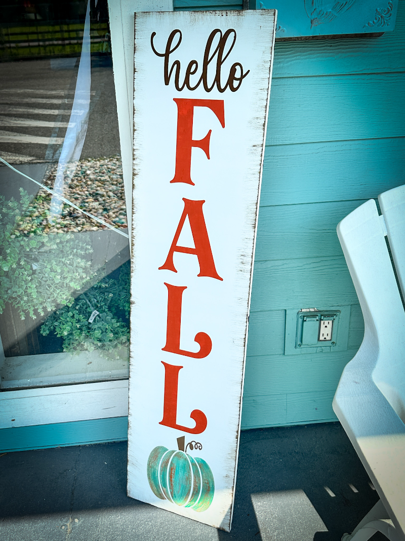 A vertical wooden plank sign with the text 'hello FALL' painted in white and red, featuring a large pumpkin graphic at the bottom.