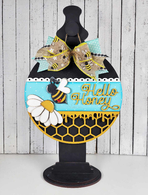 A round door hanger with a bee and honeycomb design, featuring the text 'Hello Honey' and decorative elements such as a mask and additional bees.