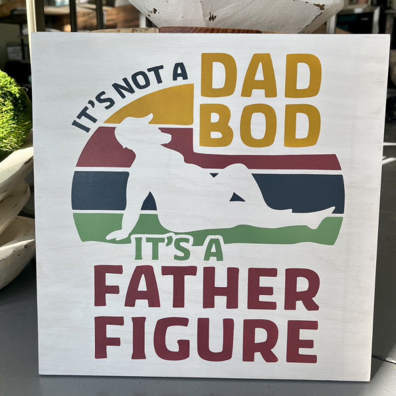 A square decorative sign with the text 'It's not a DAD BOD, IT'S A FATHER FIGURE' and a silhouette of a man in a relaxed pose with sunglasses.