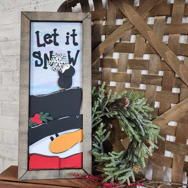 A 3D plank-style framed decoration featuring a snowman with a penguin holding a snowflake, placed on a wooden table with a wreath and pine branches in the background.