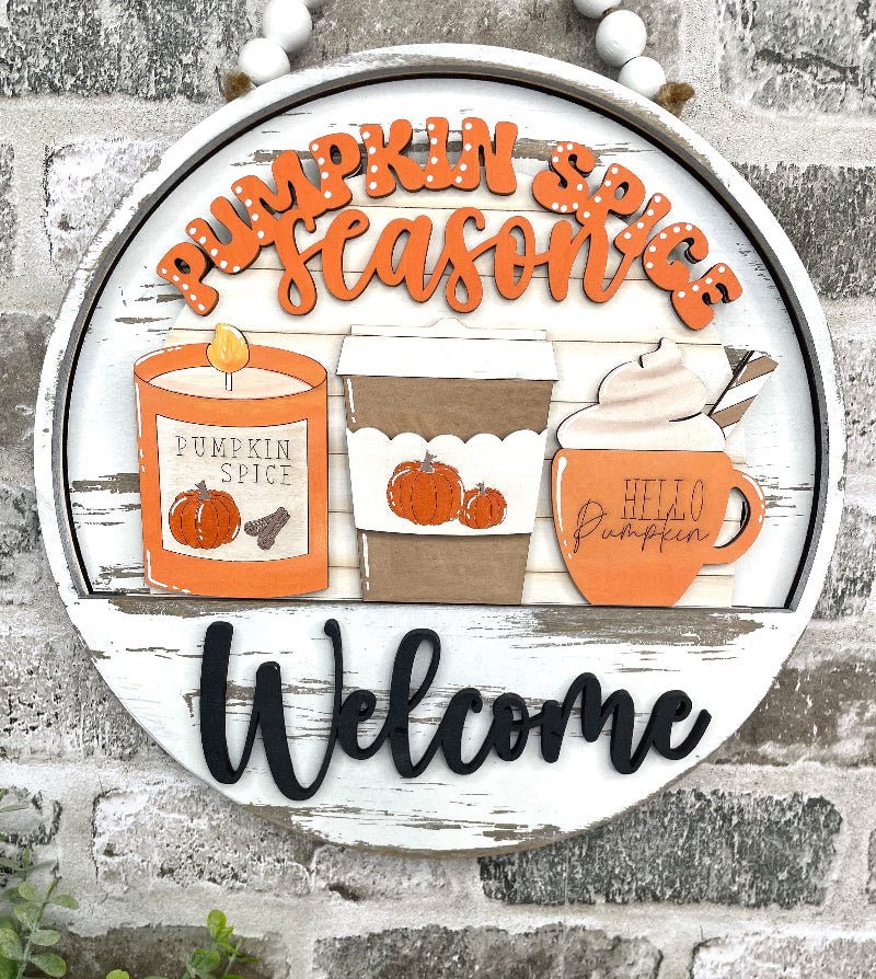 A round distressed white wooden sign with layered autumn decorations, including orange script text reading 'Pumpkin Spice Season,' illustrated pumpkin spice candle, coffee cup, and mug, and black 'Welcome' lettering at the bottom, displayed against a gray brick wall.