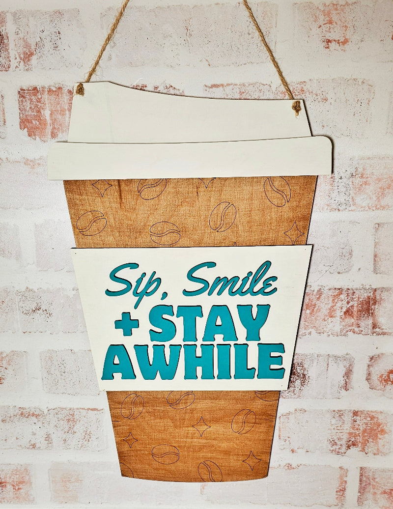 Decorative sign shaped like a coffee cup with 'Sip, Smile & Stay Awhile' text on a brick wall background.