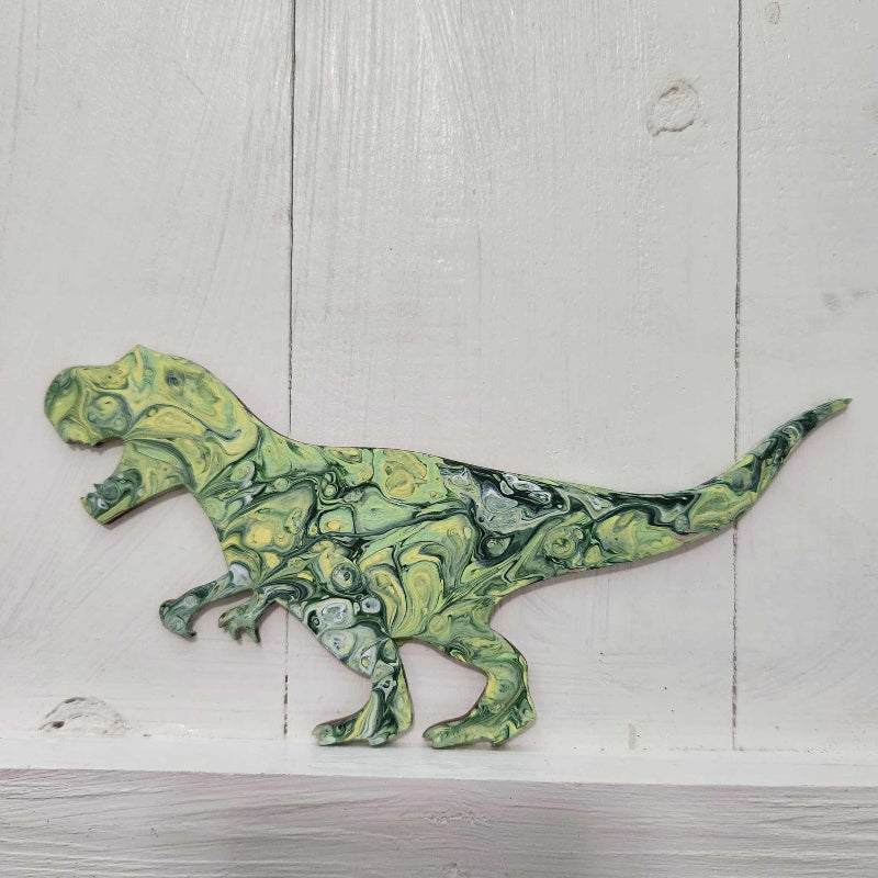 A wooden cutout in the shape of a T-Rex dinosaur with a green and yellow paint pour design on it, displayed against a white wooden background.