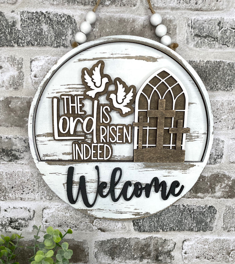 A round sign with a drop in insert that says The Lord is Risen indeed painted in a farmhouse white and brown finish.