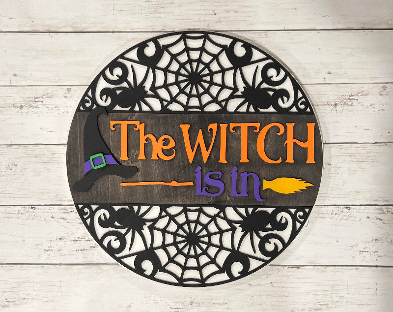 A 3D round door hanger with a witch theme, featuring a witch's hat and a cauldron, set against a wooden background with a spider web design.