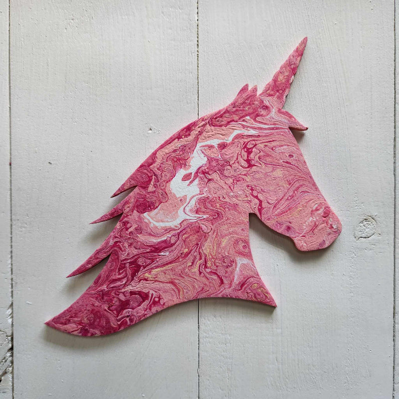 A wooden cutout in the shape of a unicorn head with a pink and red marbled paint pour design.