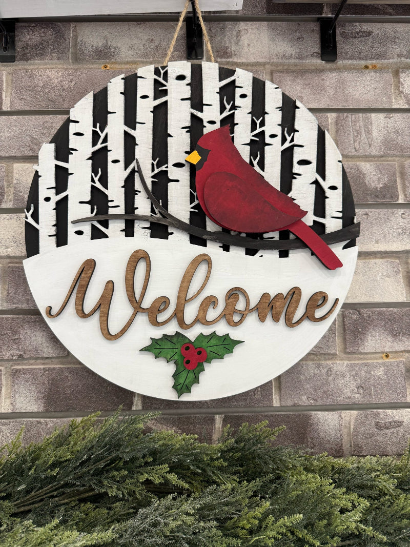 Decorative sign with a cardinal, 'Welcome' text, and holly design on a brick wall.