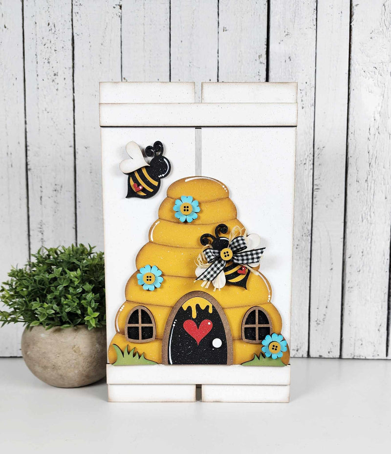 A decorative bee hive pallet sign with a bee and floral accents, displayed on a white background with a plant to the side.