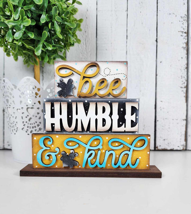 A decorative word block set with the words 'Bee Humble & Kind' displayed on a wooden stand, featuring a yellow background with bee motifs.