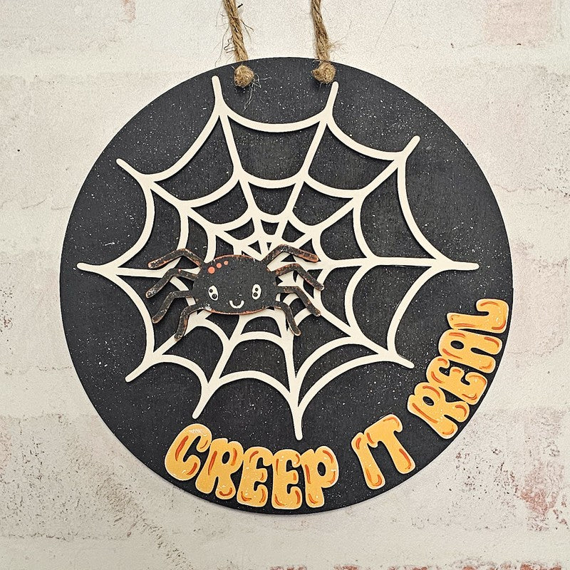 A round black Halloween wall hanging with a white spiderweb design, a small black spider in the center, and the phrase “CREEP IT REAL” in orange lettering, suspended by twine against a light textured wall.