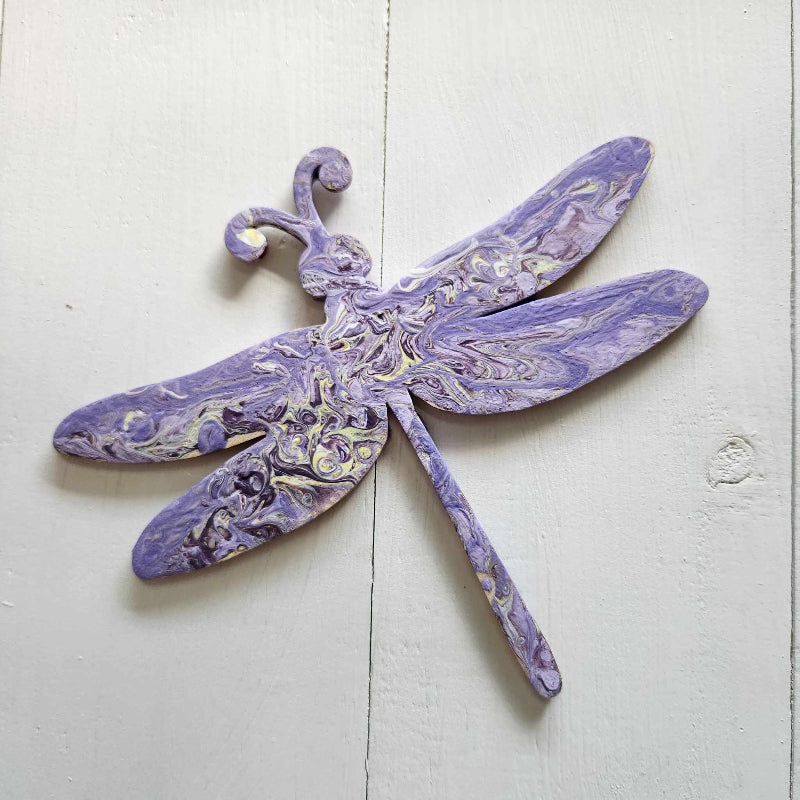 A wooden cutout in the shape of a dragonfly, with a purple paint pour design over its body.