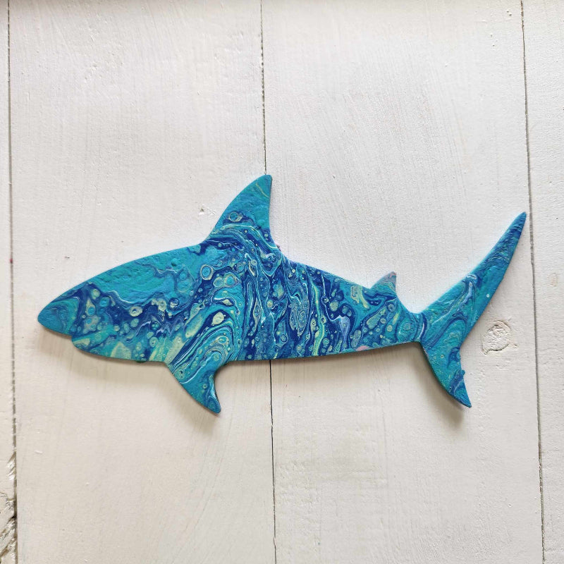 A wooden cutout in the shape of a shark with a blue paint pour design on it, placed against a white wooden background.