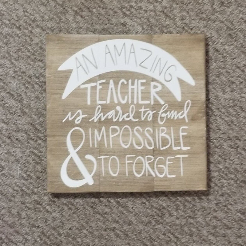 A square wooden artwork with the text 'An Amazing Teacher is hard to Find' in a whitewashed black font, placed on a floor.