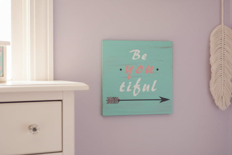 Decorative sign with 'Be you tiful' text on a wall next to a white dresser.
