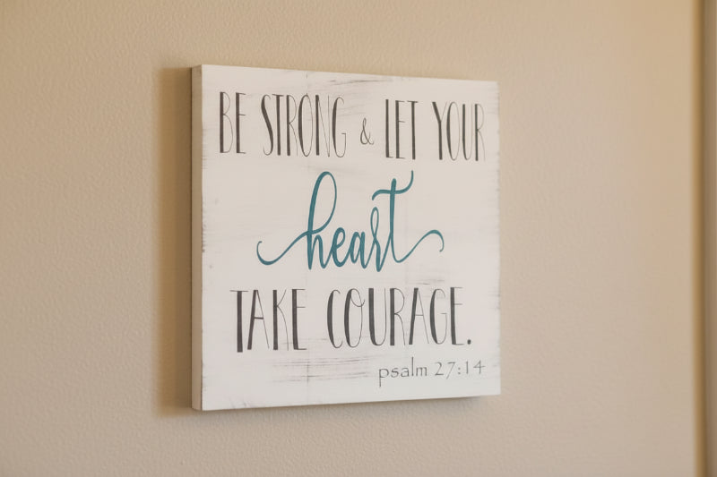 A square-shaped wall art with a wooden background featuring the text 'BE STRONG & LET YOUR HEART TAKE COURAGE' from Psalms 27:14 in a black and teal font.