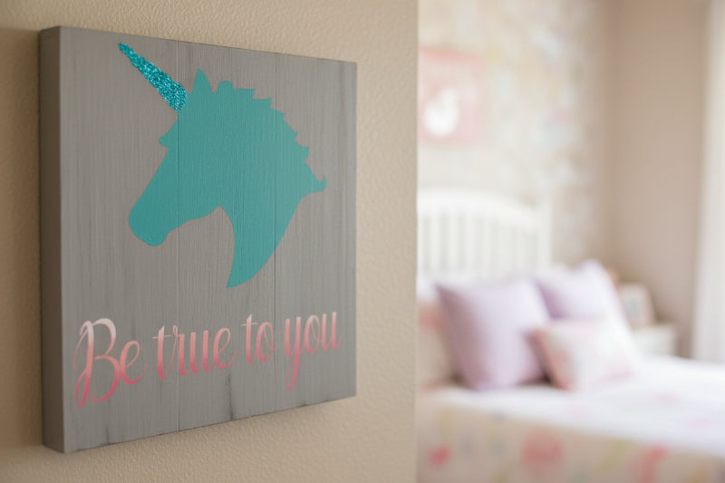 Decorative wall art with a teal unicorn head and 'Be true to you' text on a gray background, placed in a room.