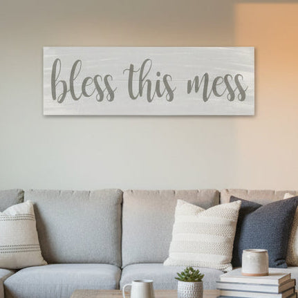 A white wooden plank sign with the phrase 'BLESS THIS MESS' written in black cursive text.
