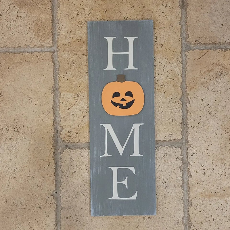 A vertical plank sign with a gray background, the word 'Home' in large white letters, and a small orange pumpkin graphic below the text.