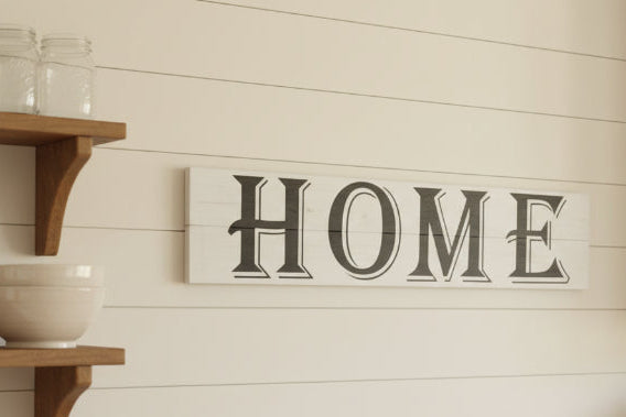 A wooden plank design sign with the word 'HOME' written in bold black letters, placed on a brown sofa.