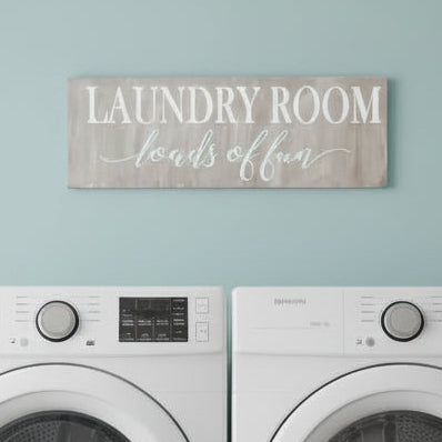 A plank design laundry room sign with the text 'LAUNDRY ROOM LOADS OF FUN' in a gray font on a simulated wooden background.