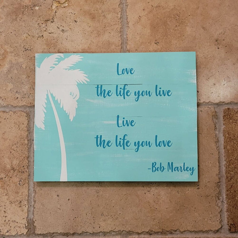 A rectangle-shaped artwork with a tropical design featuring palm trees and the text 'LOVE THE LIFE YOU LIVE AND LIVE THE LIFE YOU LOVE' by Bob Marley, on a teal background.