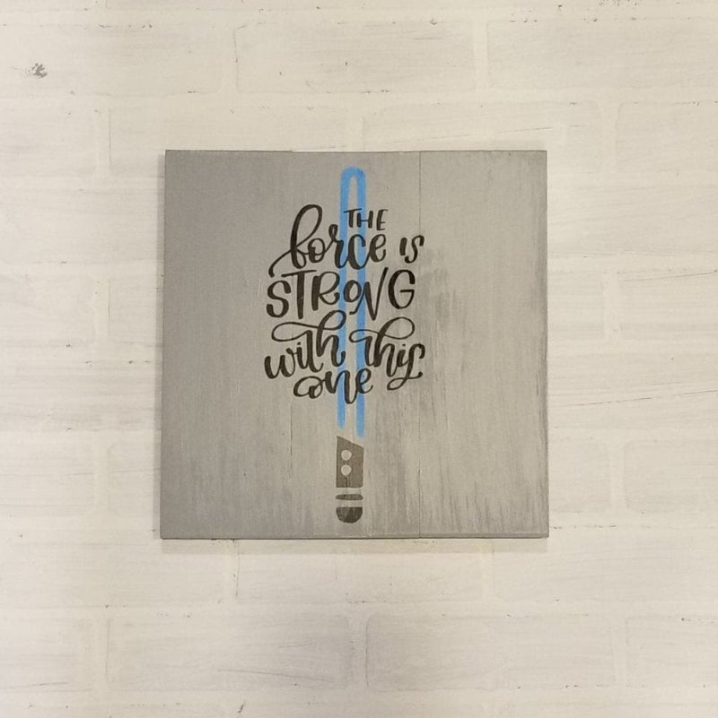 A square-shaped wall decor featuring the text 'THE FORCE IS STRONG WITH THIS ONE' in a stylized font, with a representation of a lightsaber on the right side of the text.