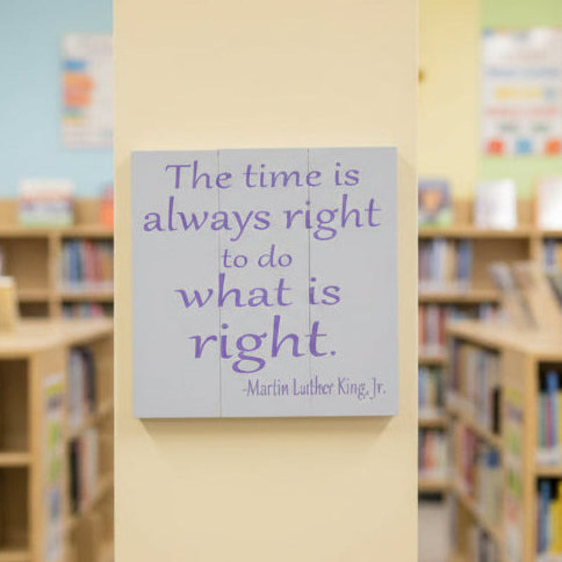 A square-shaped wall art featuring a quote by Martin Luther King Jr. with a purple-colored text on a gray background, placed on a tiled floor.