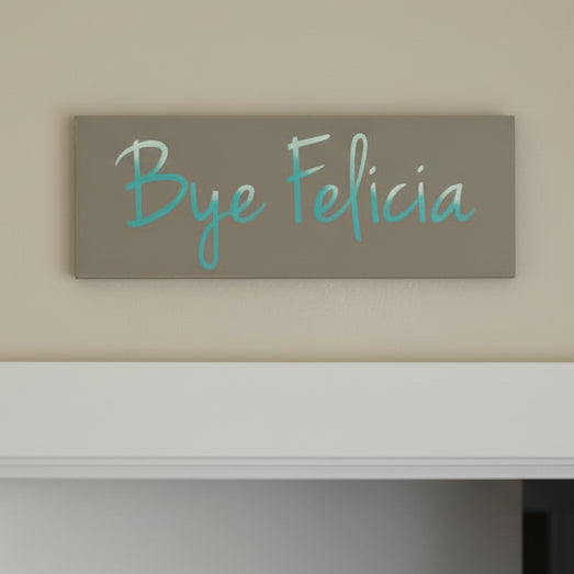 A humorous plank design sign with the text 'Bye Felicia' in a stylized font, placed on a wooden surface.