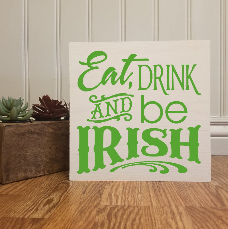 A square design decorative plaque with the text 'EAT DRINK AND BE IRISH' in bold green letters on a white background, placed against a wooden backdrop with small plants on either side.