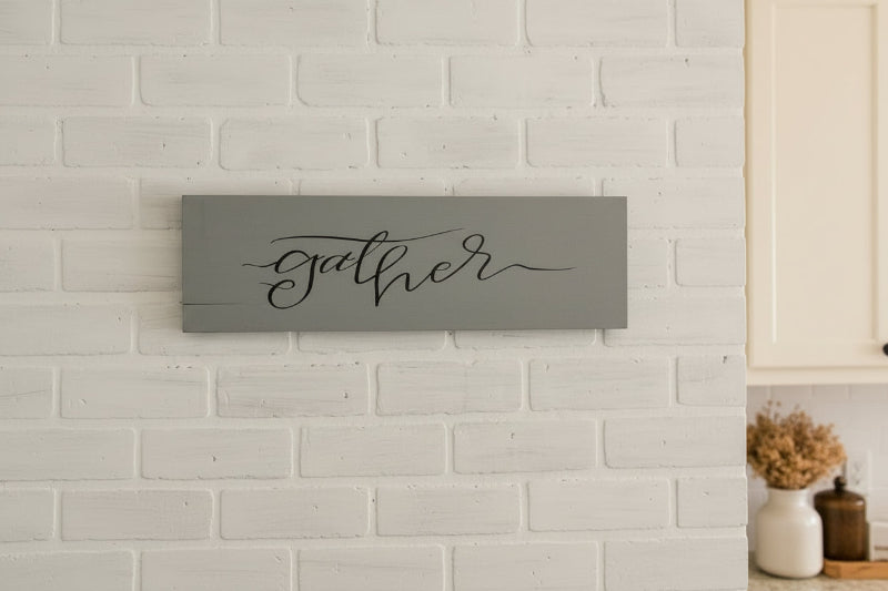 A gray-colored plank design sign with the word 'GATHER' written in a cursive script, mounted on a white brick wall.