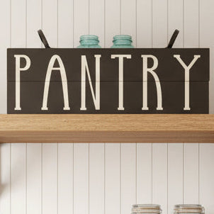A black plank design sign with the word 'Pantry' written in white. It is displayed above a small blue jar and coasters.