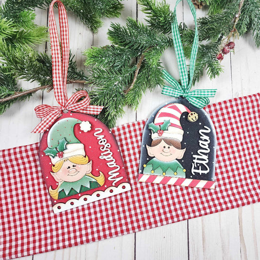 Two Christmas-themed ornaments with names on a checkered cloth and wooden surface.