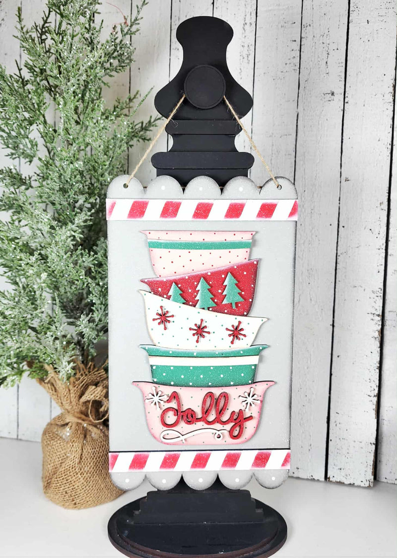 Decorative Christmas sign with colorful bowls and 'Jolly' text on a white background.