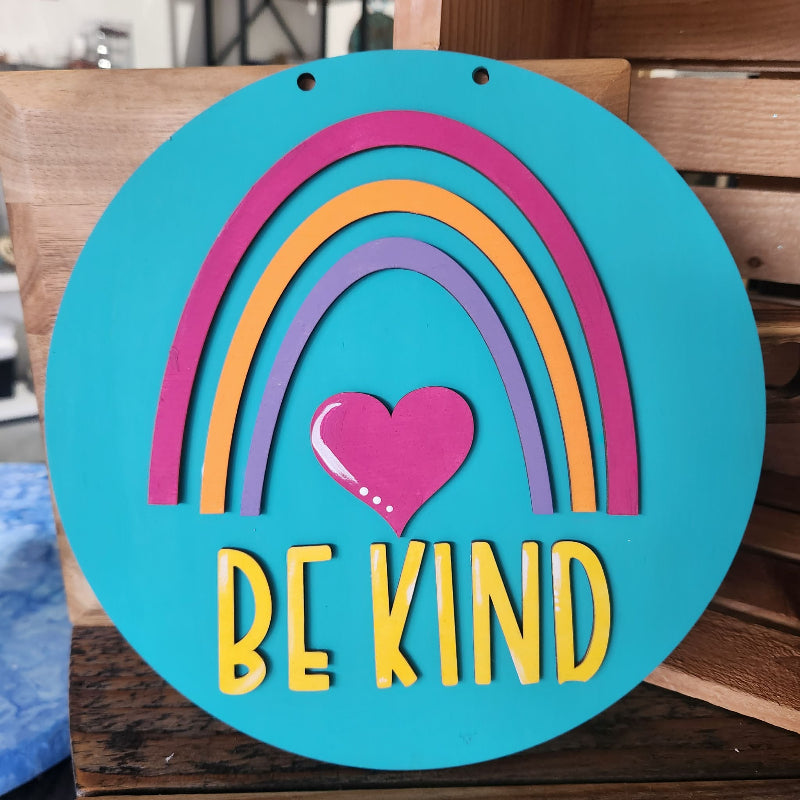 Decorative sign with rainbow and heart design on a teal background, featuring the text 'BE KIND'.