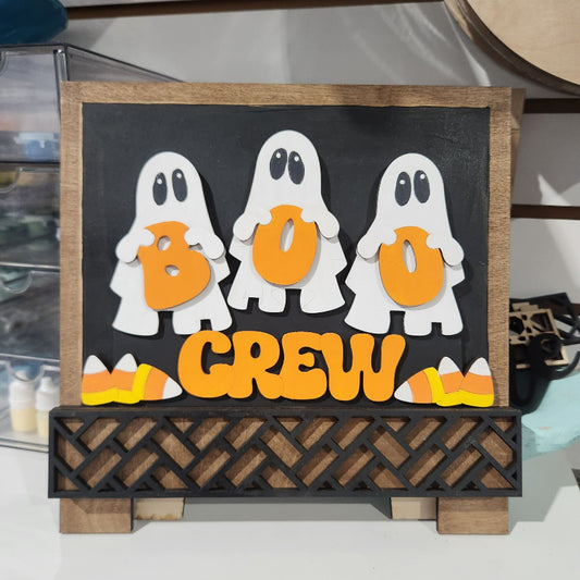 A wooden Halloween-themed sign reading 'BOO CREW' with three white ghosts holding orange letters and candy corn accents, displayed on a shelf.