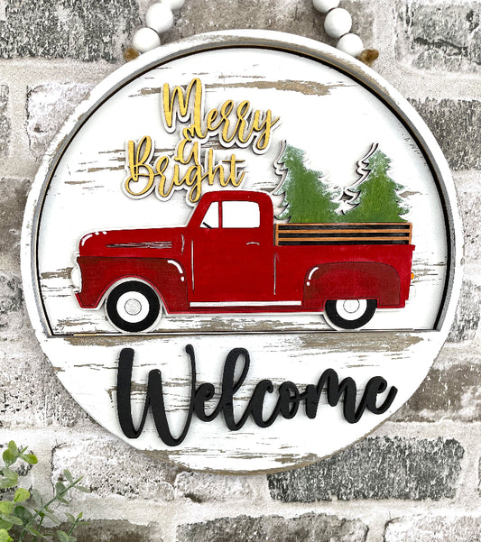 A red truck with trees in the back, the words Merry & Bright written in yellow placed on a round 10.5 in door hanger that says Welcome.