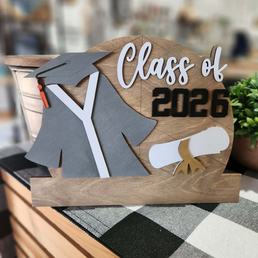 Decorative sign with 'Class of 2026' text, graduation cap, gown, and diploma on a wooden background.