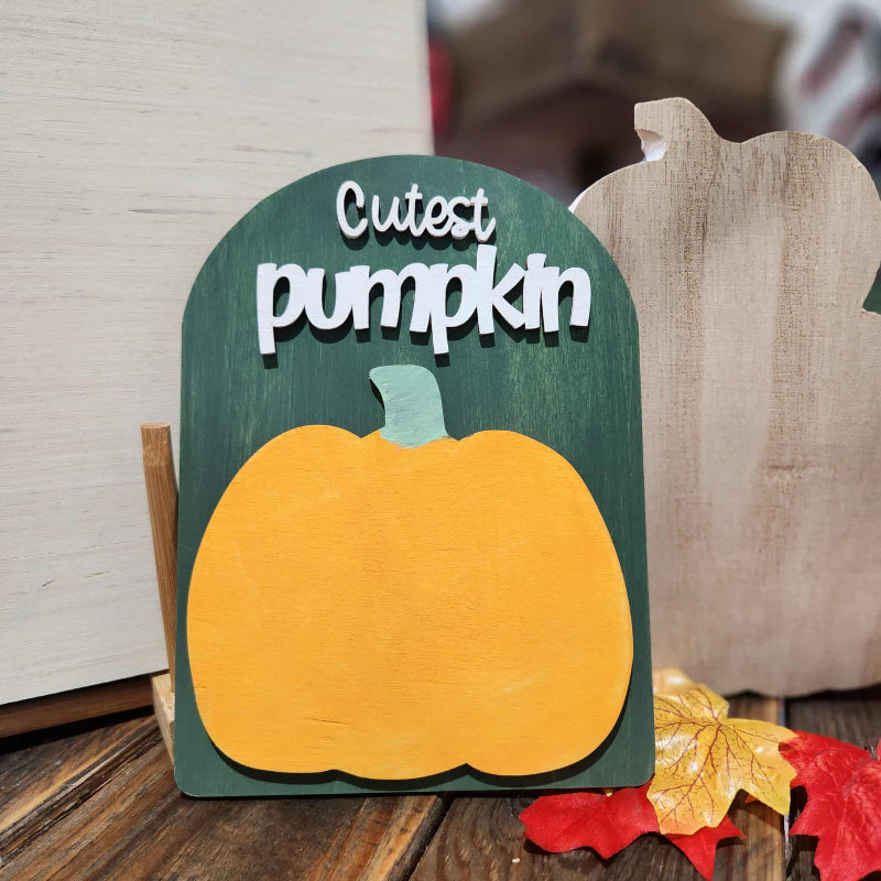 Decorative sign with a pumpkin and 'Cutest Pumpkin' text on a wooden surface with leaves.