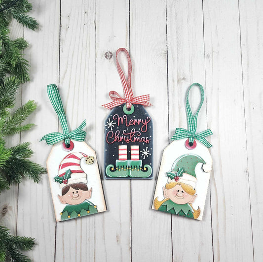 Three Christmas-themed tags with elves on a wooden surface.