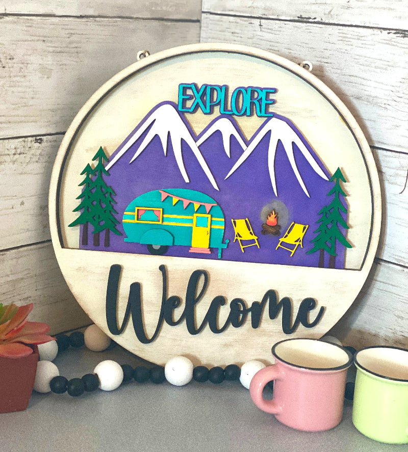 Alt text: Explore camping interchangeable wooden Welcome sign insert with purple mountains, evergreen trees, teal camper, campfire and yellow camp chairs.