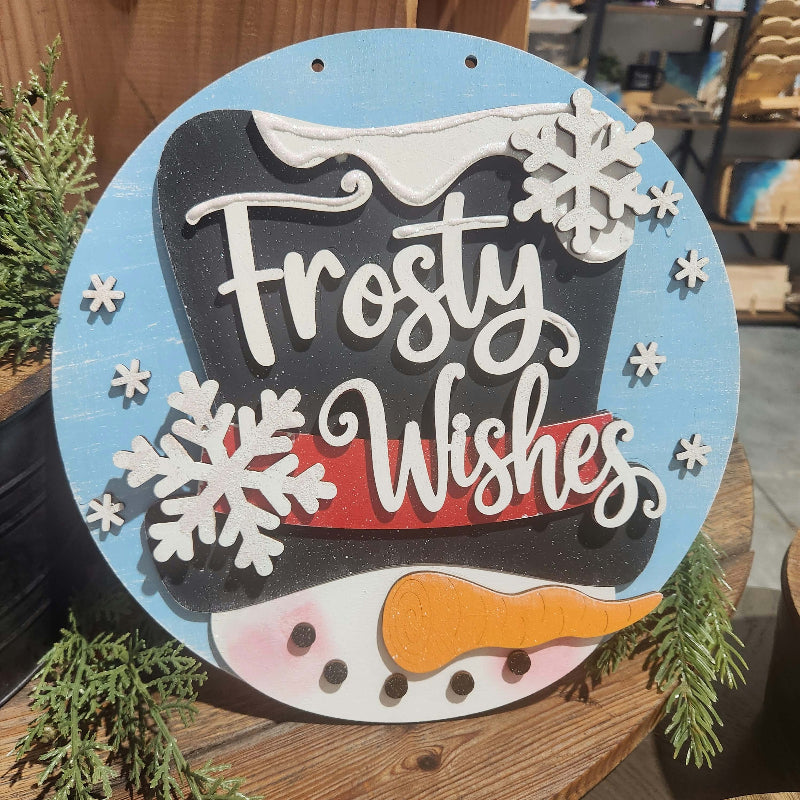 : Frosty Wishes round layered wooden snowman sign featuring a black top hat, white snowflakes, orange carrot nose, and blue winter background.