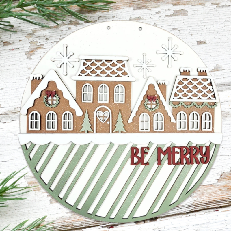 Decorative Christmas ornament with gingerbread house design and 'Be Merry' text on a wooden surface.
