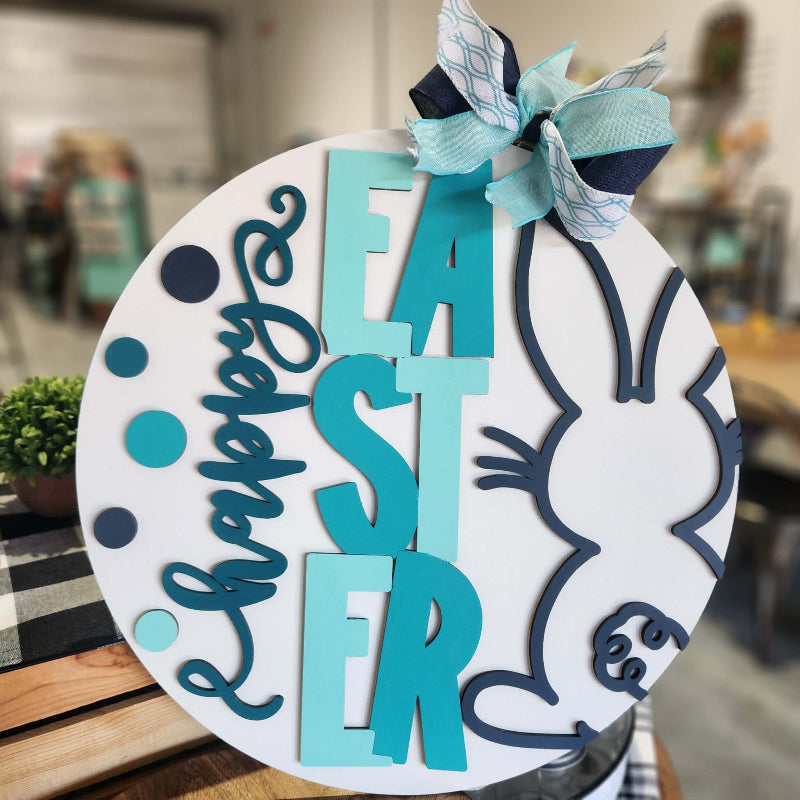 Decorative Easter sign with blue and white text and a bunny design, placed on a wooden surface.